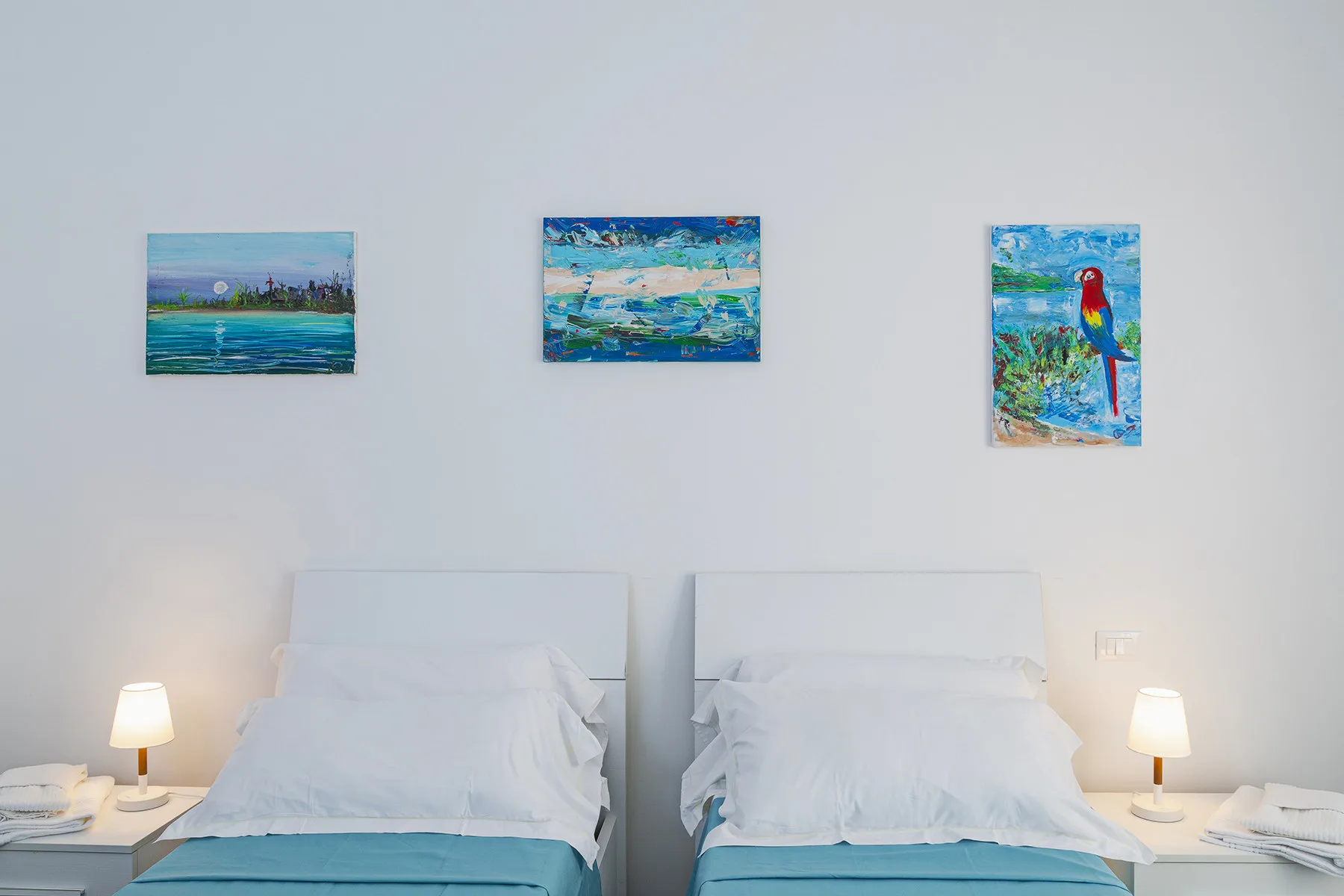 A bright bedroom has two beds with white bedding and colorful paintings on the wall.