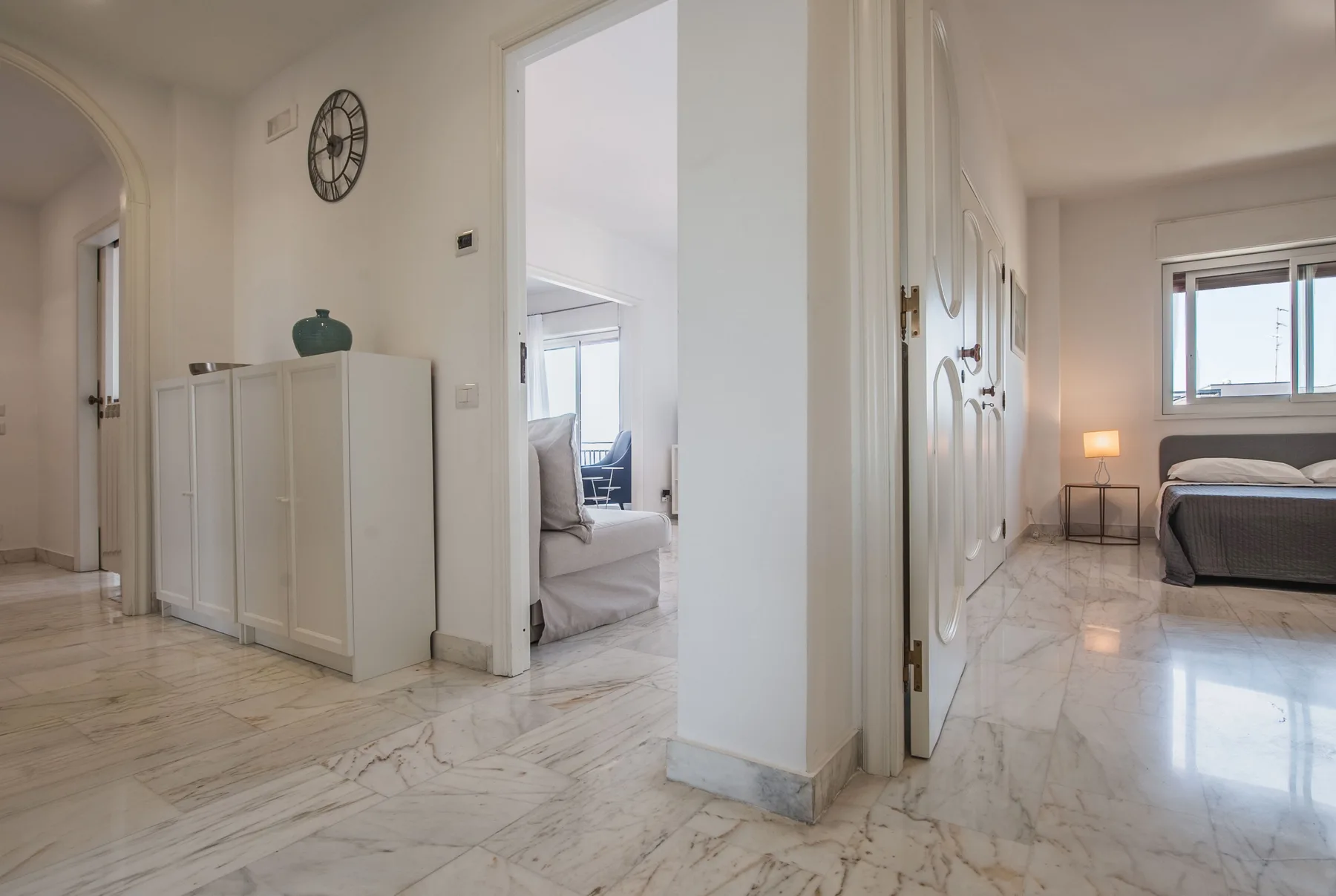 A bright interior hallway opens onto a bedroom with a bed and marble flooring.