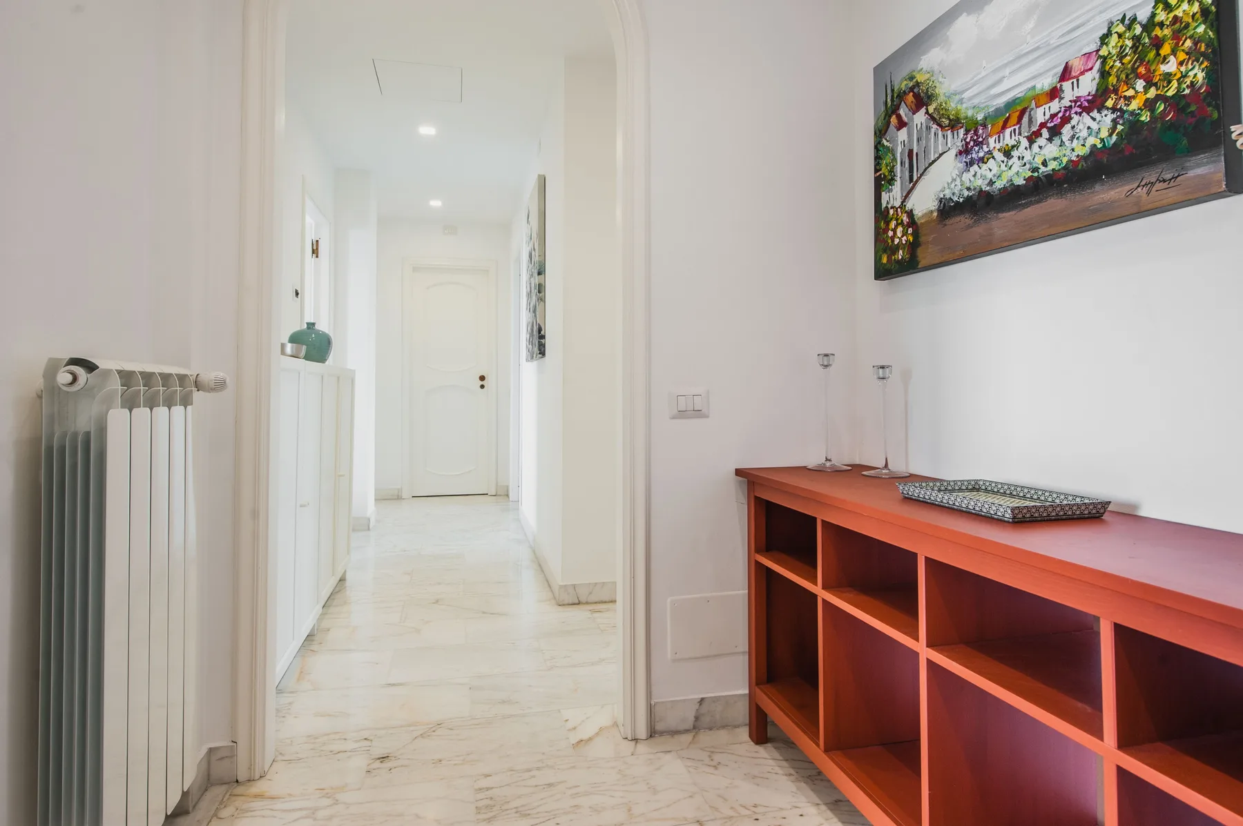 A bright hallway with a radiator, a wooden shelf, and colorful wall art.