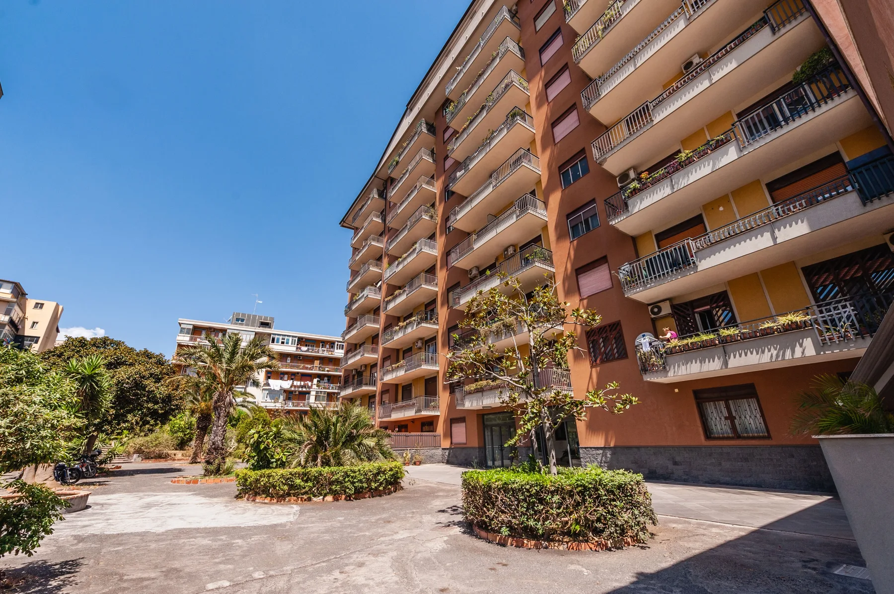 A paved courtyard with planted shrubs sits beside a tall apartment building.