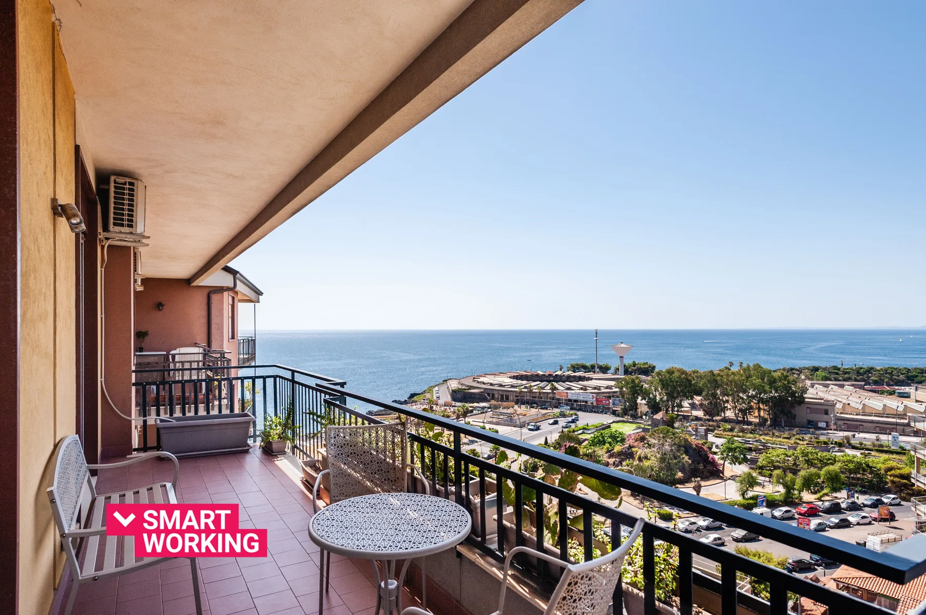 A balcony overlooks the sea, with outdoor seating and a black railing.