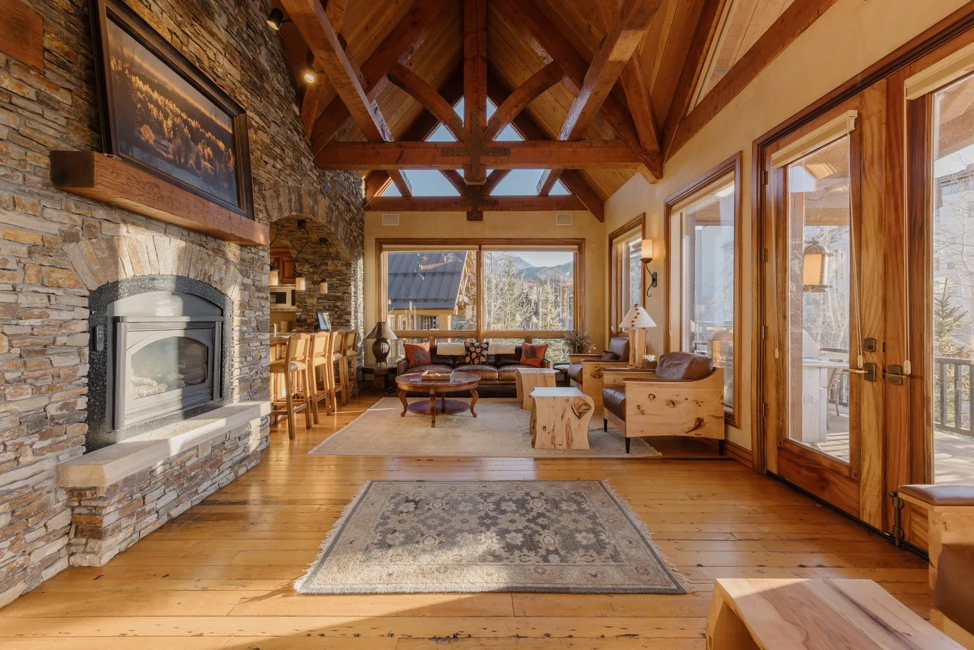 A wood-beamed living room with a stone fireplace and large windows.