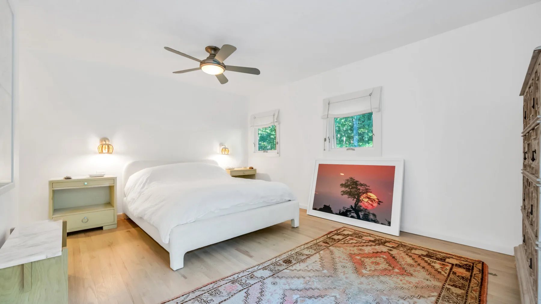Un dormitorio luminoso con una cama blanca, una alfombra colorida y dos ventanas.