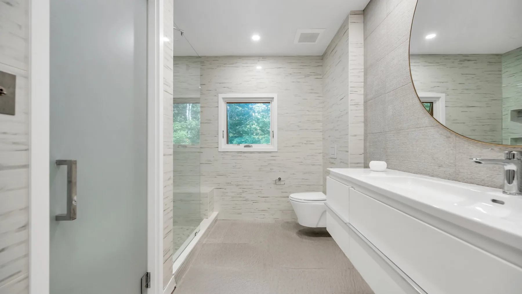 Un baño luminoso con azulejos, una ducha de vidrio, una ventana y un tocador largo.