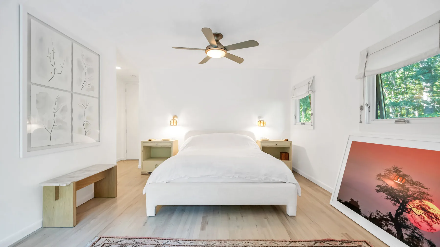 Un dormitorio luminoso con una cama blanca, muebles de madera clara y ventanas amplias.