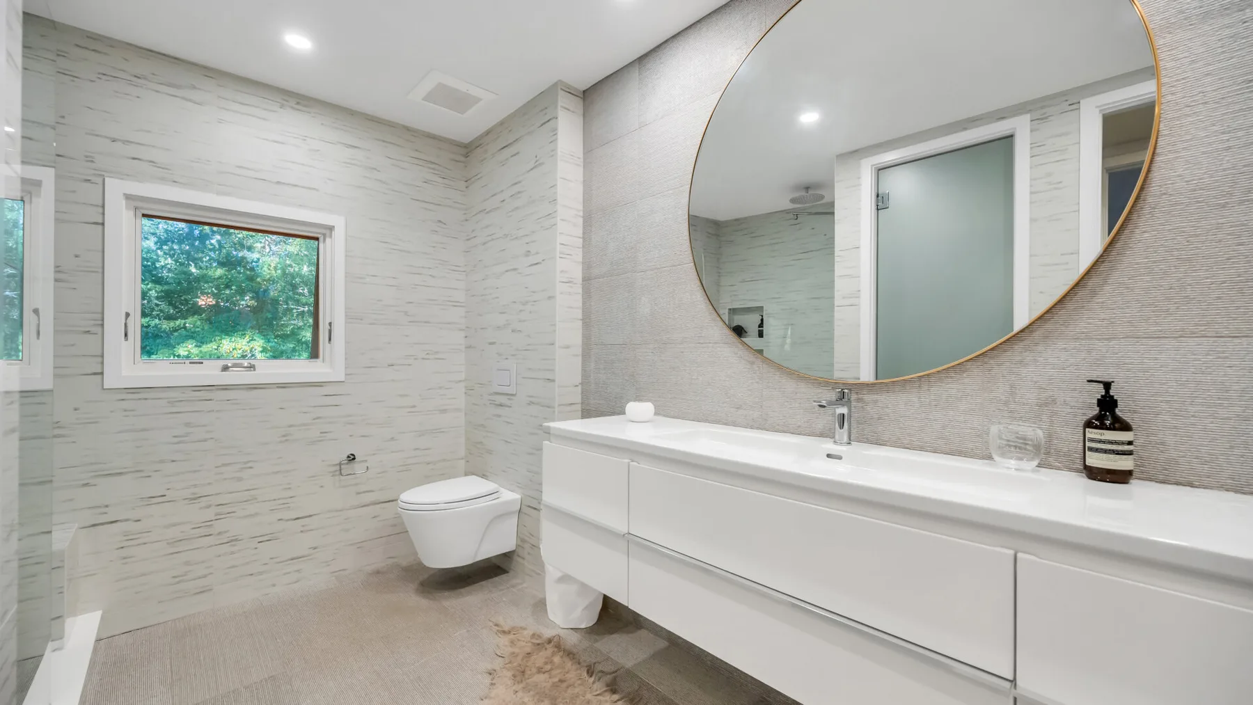 Un baño luminoso con un tocador largo, un espejo redondo y una ventana.