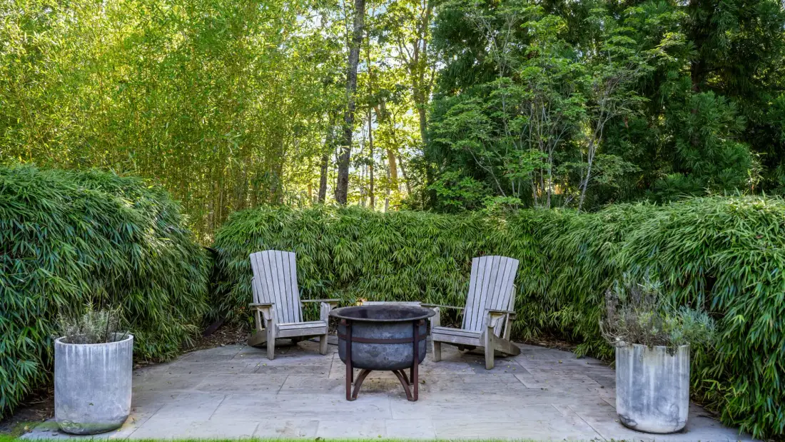 An outdoor patio with two lounge chairs around a round fire pit, surrounded by greenery.