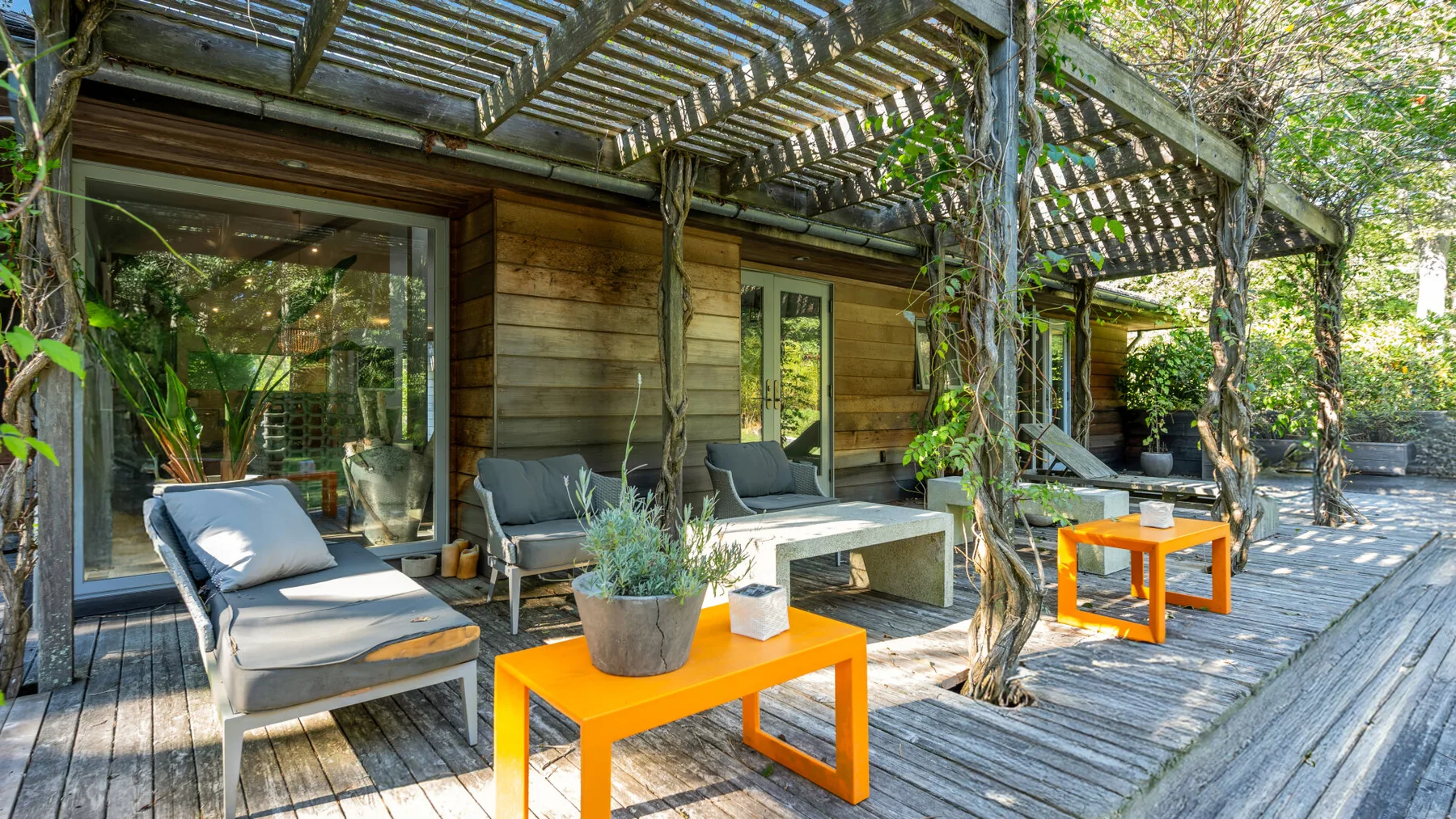 A wooden patio beneath a pergola has lounge seating and bright orange tables.