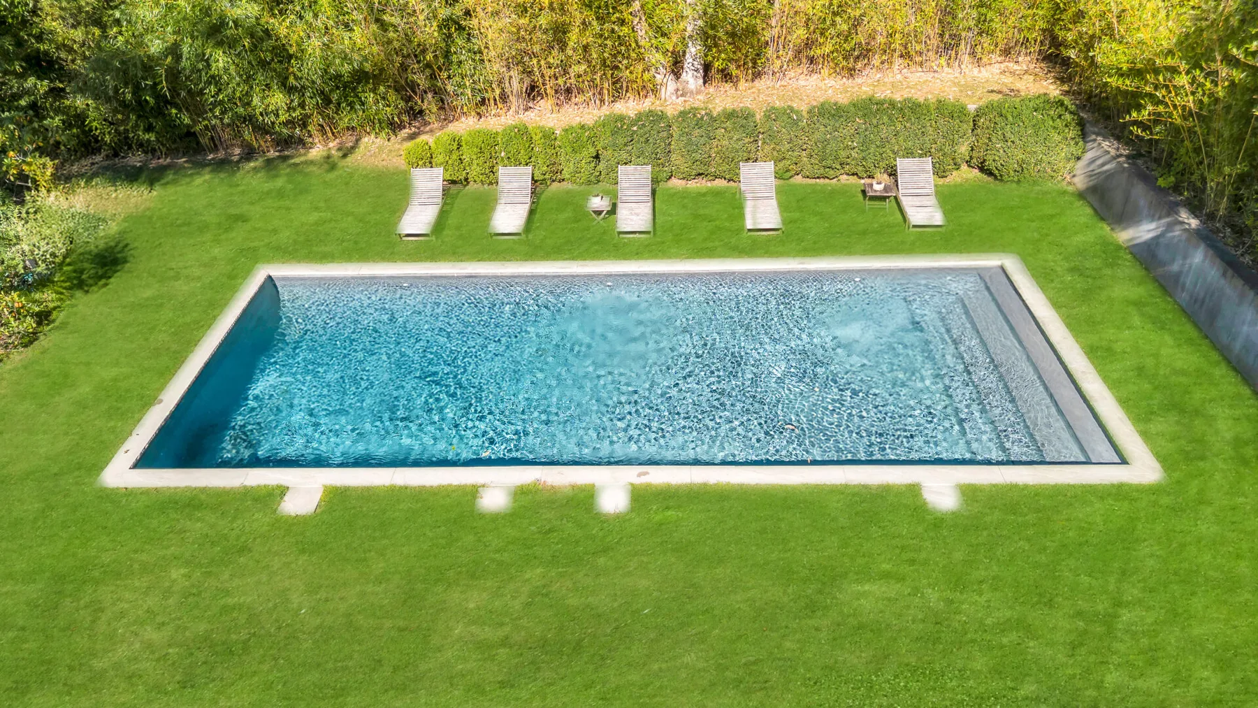 Una piscina rectangular está en un jardín con césped y tumbonas cerca.
