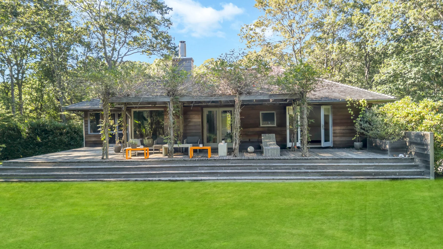 Una casa de madera da a una amplia terraza junto a un césped verde y árboles.