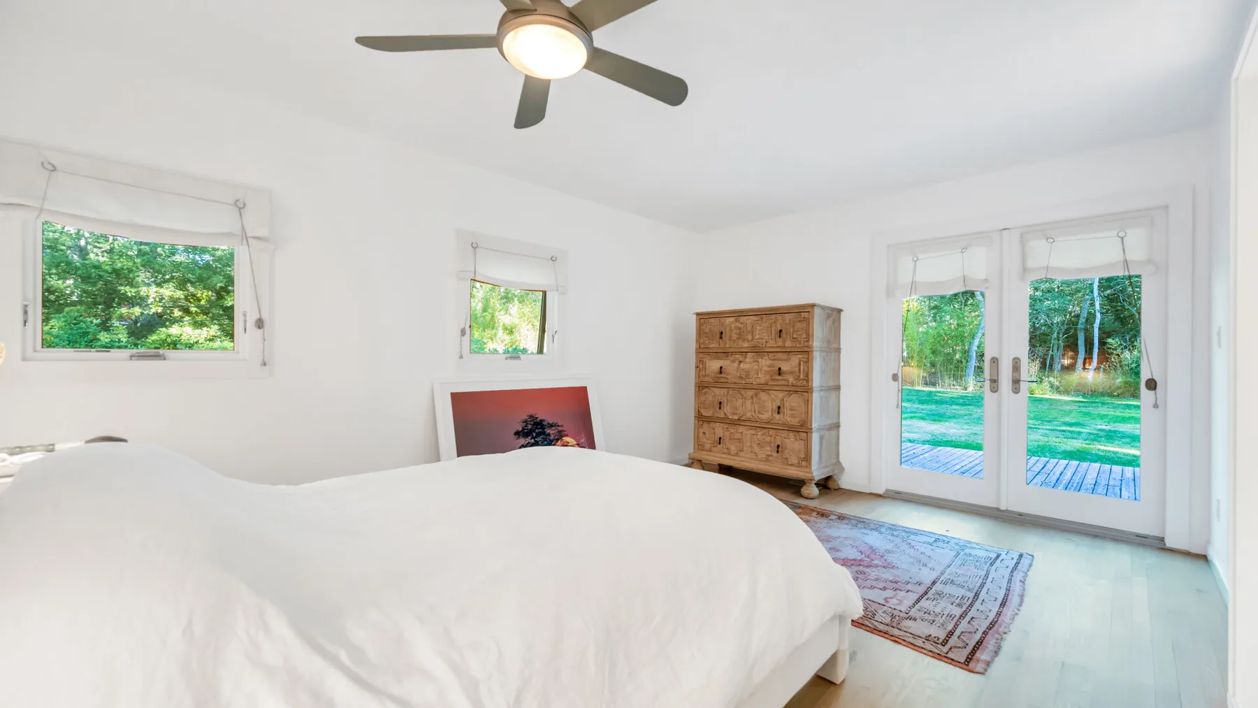 Un dormitorio luminoso con una cama blanca, ventilador de techo y puertas de vidrio hacia un patio.