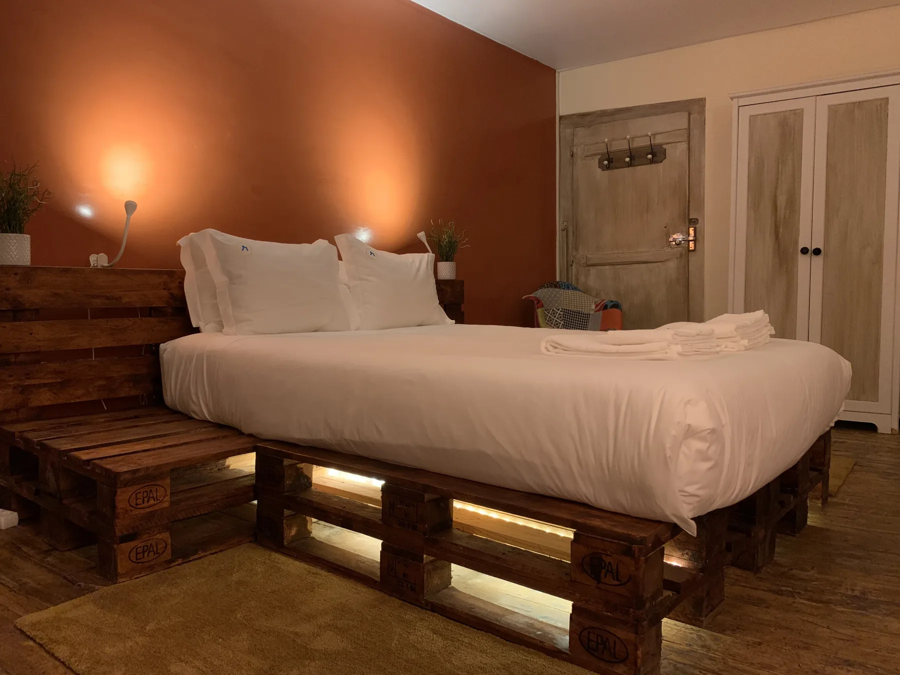 Un dormitorio con una cama blanca, una base de madera tipo palé y luz cálida en la pared.