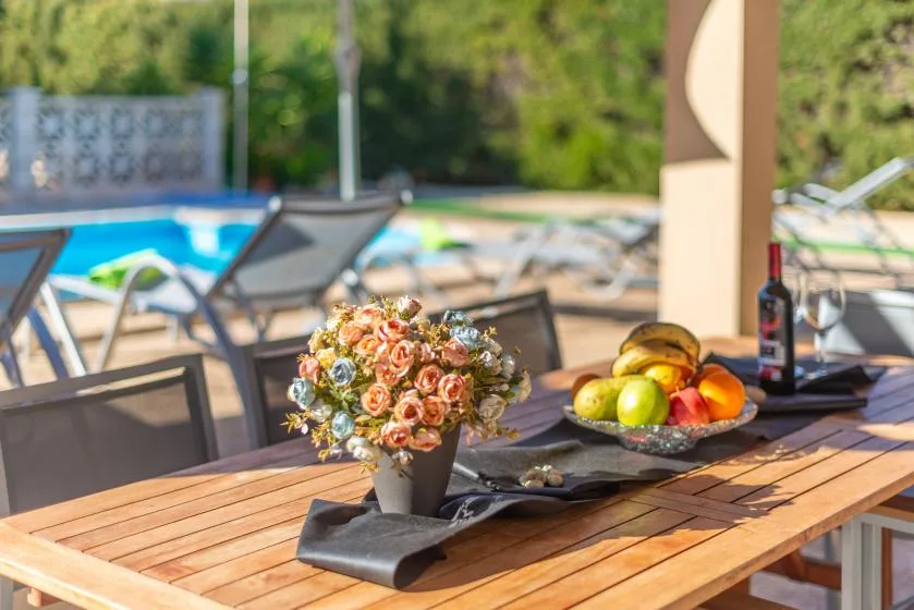 Una mesa de comedor al aire libre con flores y fruta junto a una piscina.