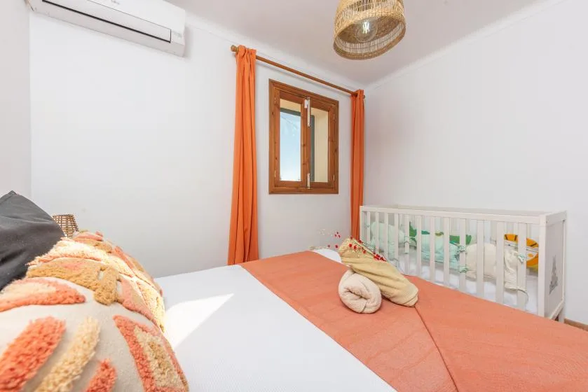 Un dormitorio con una colcha naranja, una ventana y una cuna blanca.