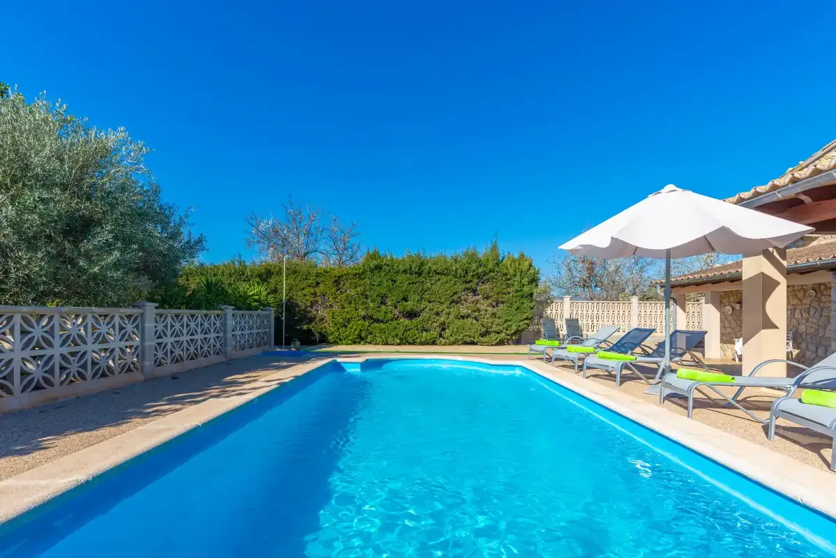 Una piscina exterior junto a un patio con tumbonas y una sombrilla.