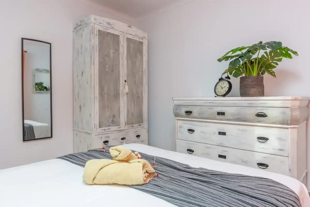 Un dormitorio luminoso con una cama y armarios y cómoda blancos envejecidos.