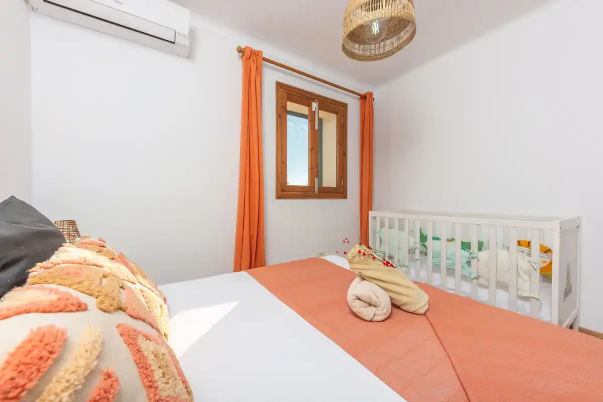 Un dormitorio con ropa de cama naranja, una ventana y una cuna blanca.