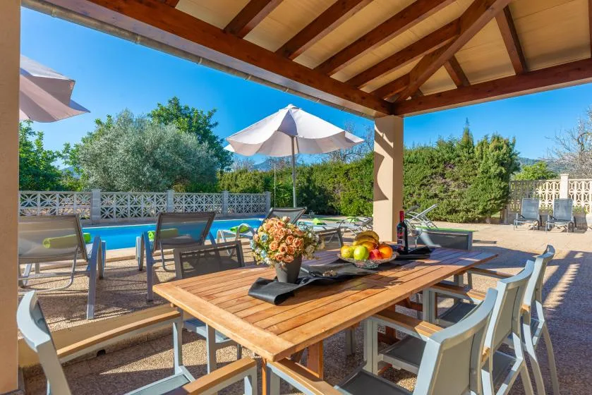Una zona de comedor al aire libre con sombra da a una piscina y un jardín frondoso.