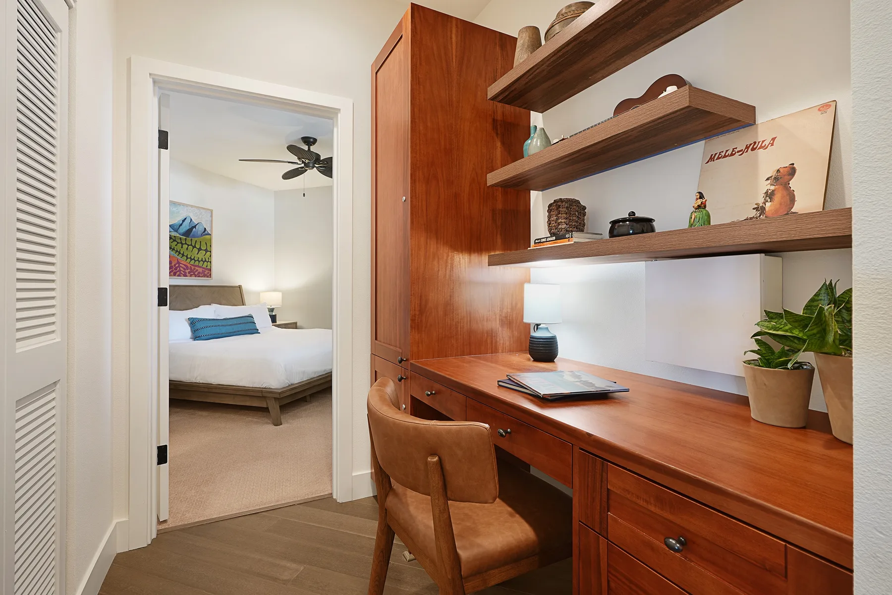 Un escritorio y estantes de madera ocupan un rincón de estudio junto a la puerta abierta de un dormitorio.