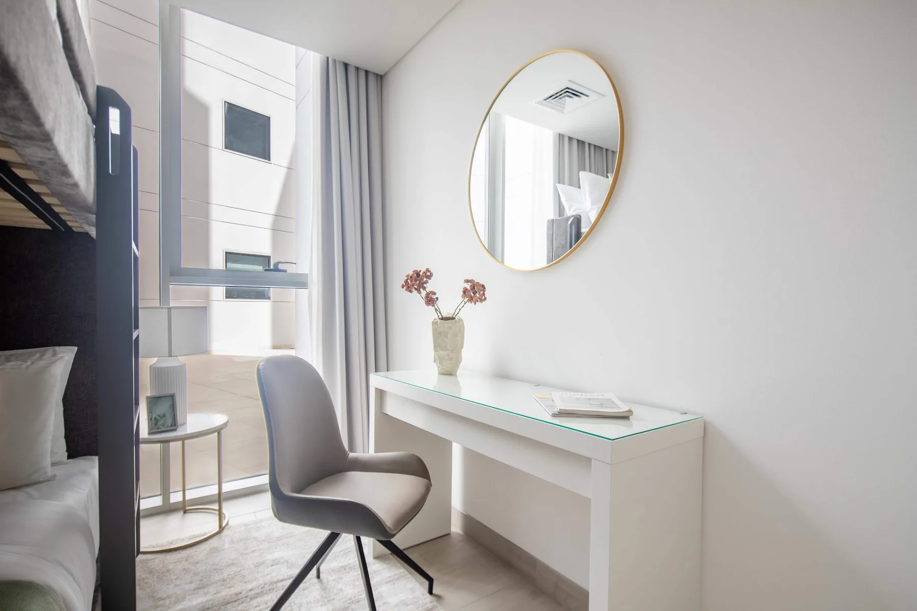 A bright bedroom corner with a desk, chair, and round wall mirror.