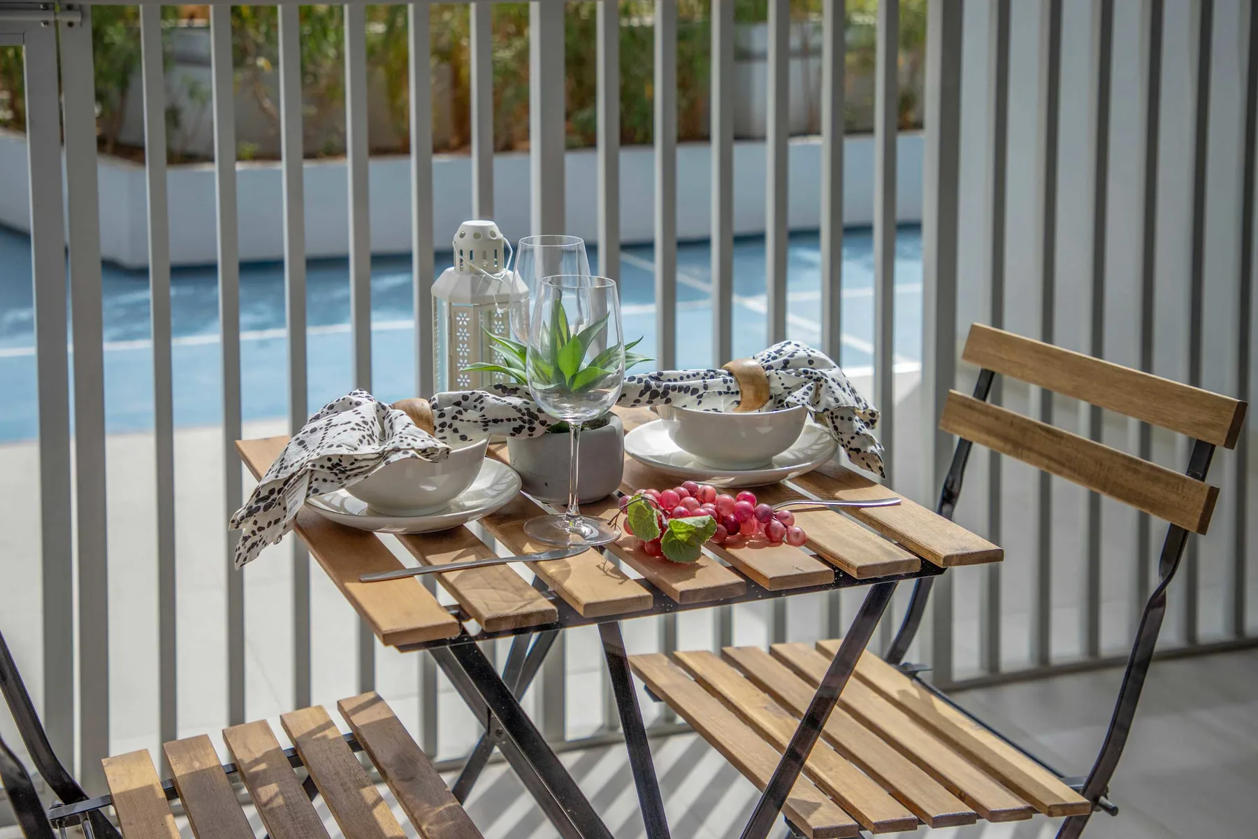 A small outdoor dining table is set with dishes and fruit beside a swimming pool.