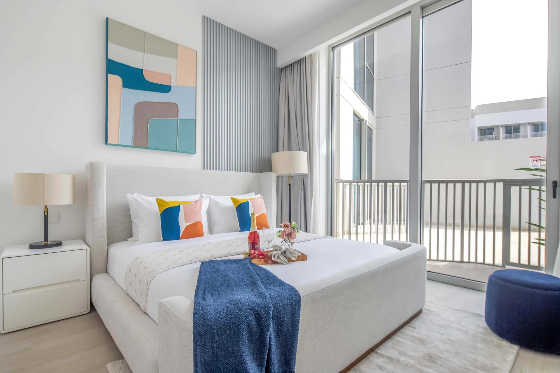 A bright bedroom with a white bed beside large windows opening onto a balcony.