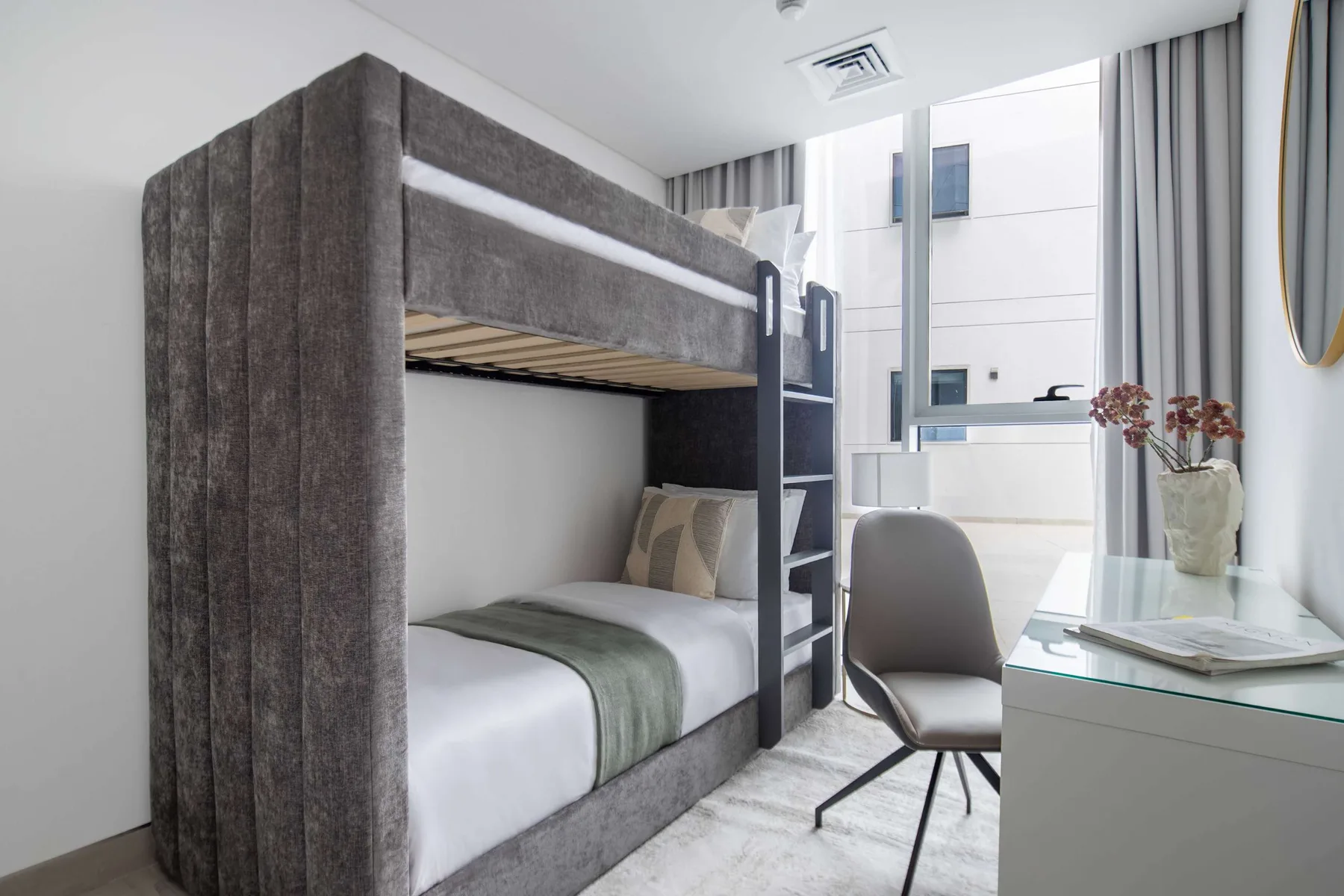 A bright bedroom with a gray bunk bed beside a desk and large window.