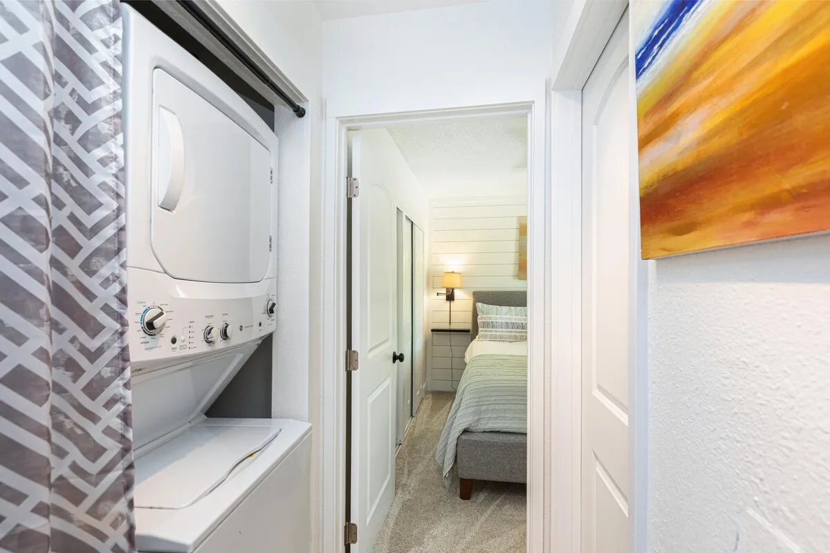 Una zona de lavandería da a un dormitorio con una cama y una lámpara de noche.