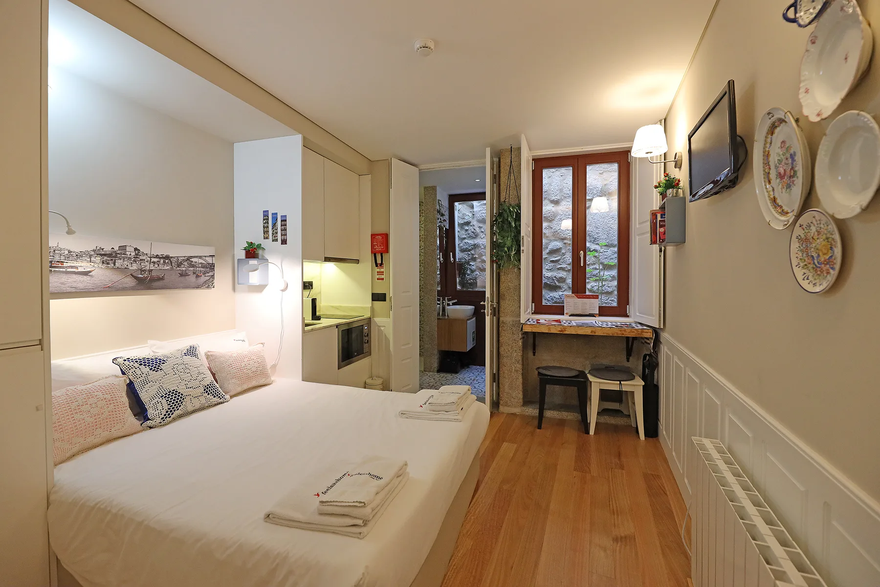 A studio room with a bed, compact kitchenette, and a window-side table.