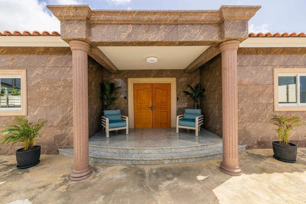 An elegant stone entrance with grand columns and welcoming seating marks the arrival to the property, framed by lush greenery and terracotta tile roofing.