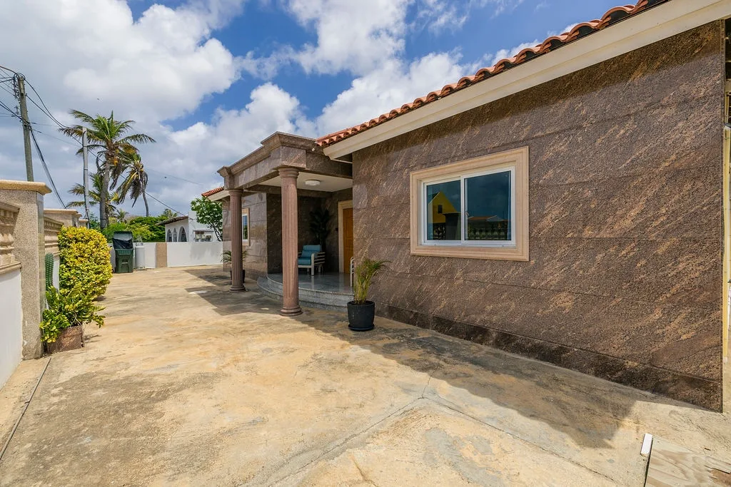 A welcoming tropical retreat with terracotta roof tiles, textured stone exterior, and columned entry opening to airy indoor-outdoor living spaces. Lush greenery and palm trees frame the property under bright island skies.