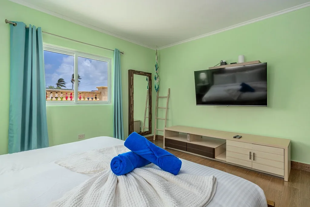 Unwind in your serene bedroom featuring calming mint-green walls, a plush bed adorned with fresh towels, and views of swaying palms and sunny skies through your window.
