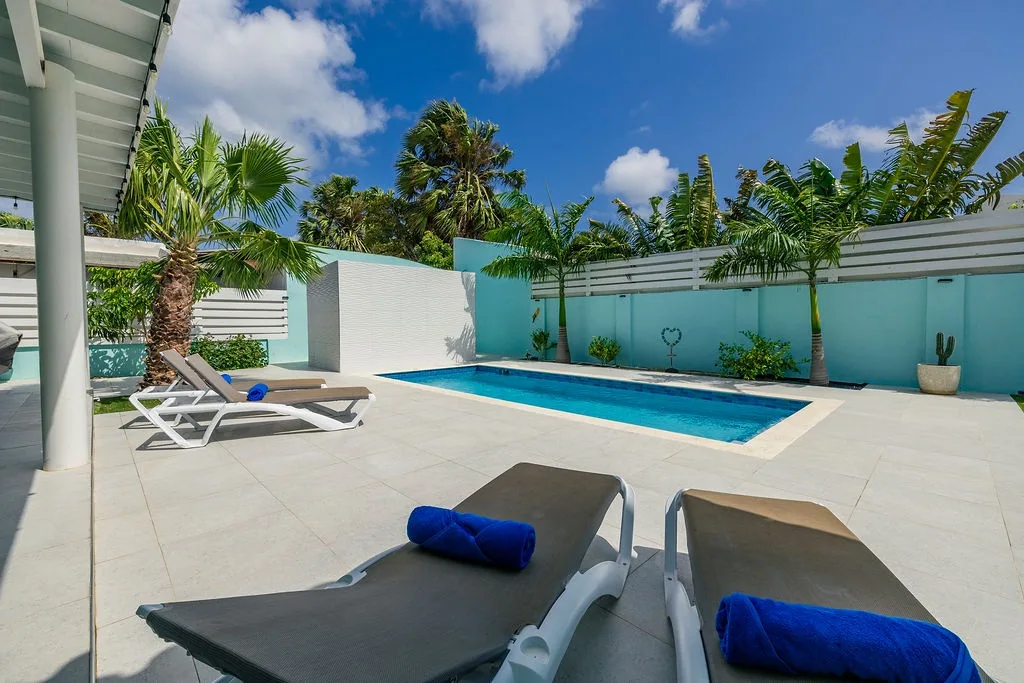 Step into your private tropical oasis, where a sparkling pool awaits beneath swaying palms and brilliant blue skies. Your lounger is ready with plush towels—perfect for soaking up the sun in complete seclusion.
