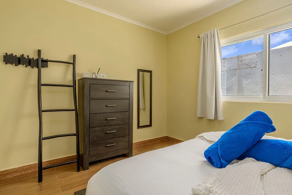 Your bright bedroom features warm yellow walls, a comfy bed with fresh towels, and plenty of storage in the tall dresser. Natural light pours in through the large window.