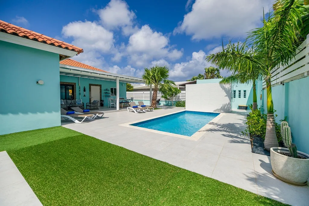 Aerial view of this inviting tropical property featuring turquoise buildings, a private pool surrounded by lush palms, and spacious outdoor relaxation areas under bright Caribbean skies.