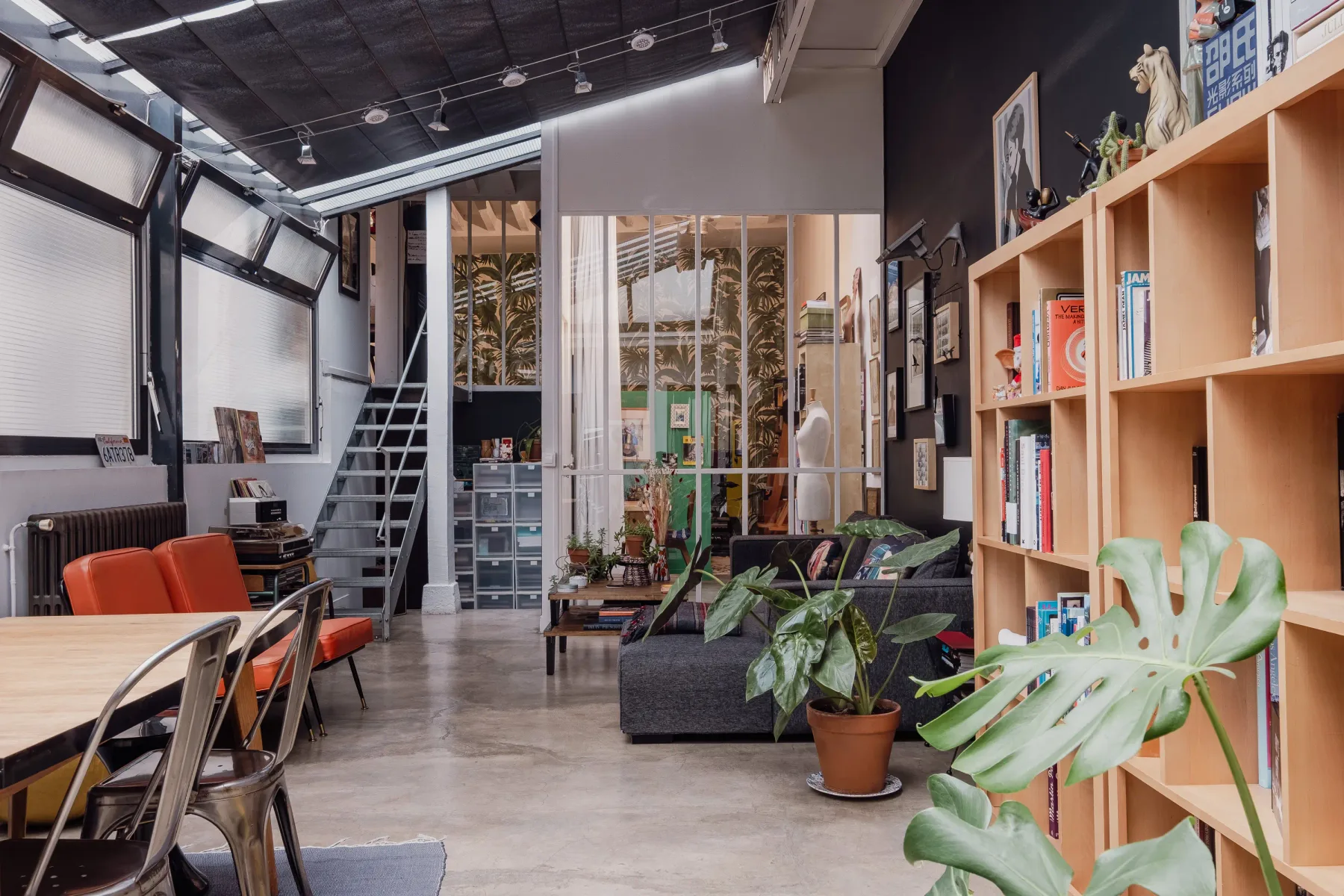 An industrial-style living room with seating, a bookshelf, and large windows.