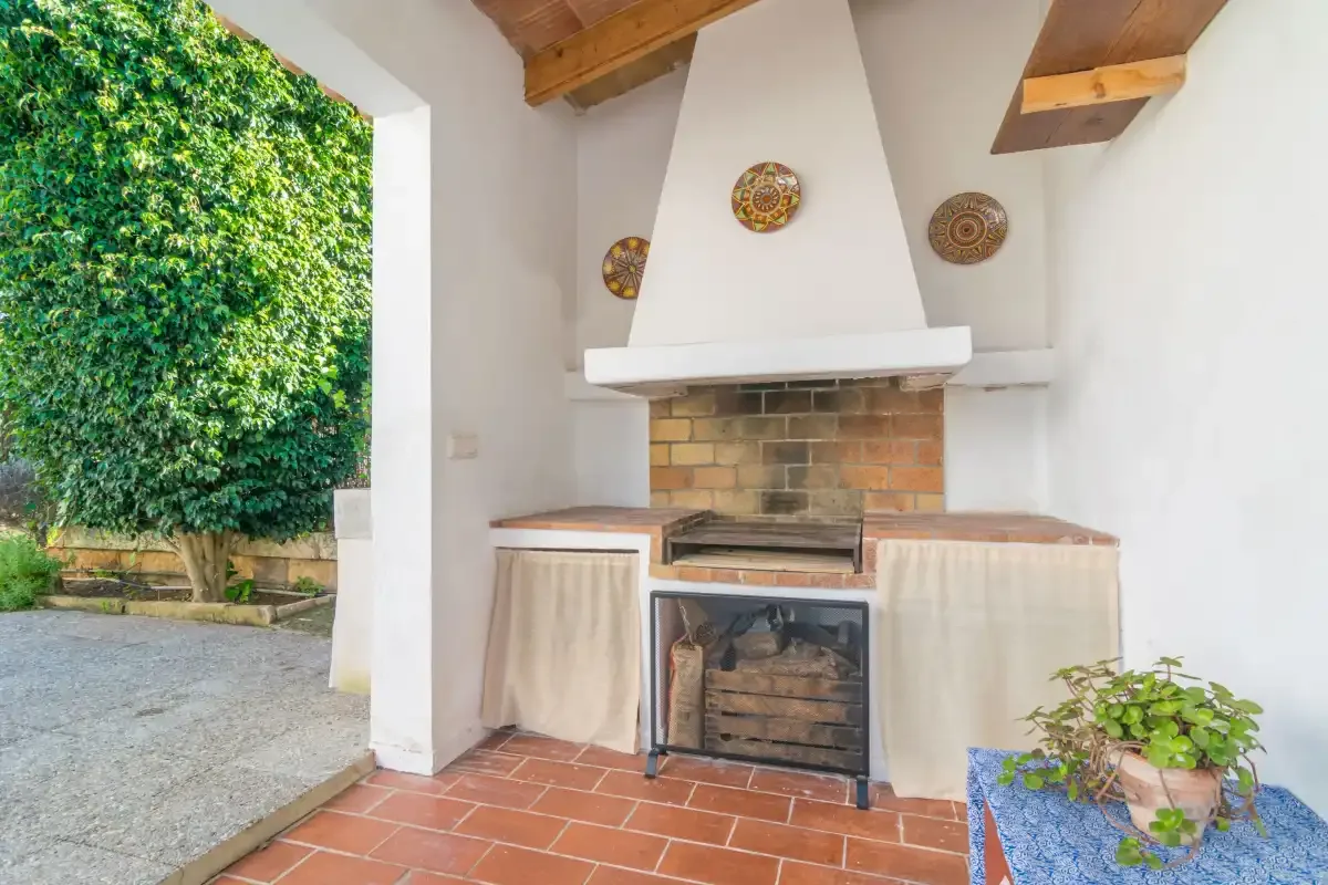 An outdoor barbecue area with a brick grill, tiled floor, and leafy greenery.