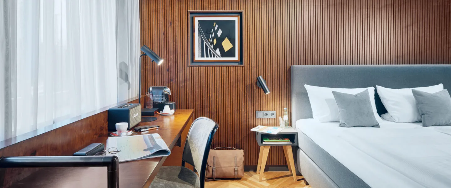 Hotel room with a bed and small desk, wood flooring and wood wall, in warm lighting.