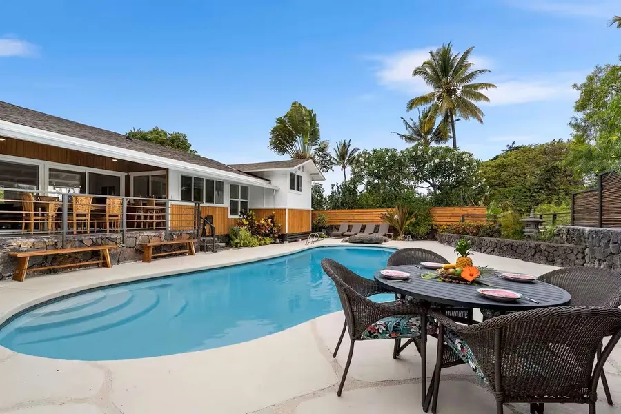 A swimming pool beside a patio dining area, with a house and palm trees in the background.