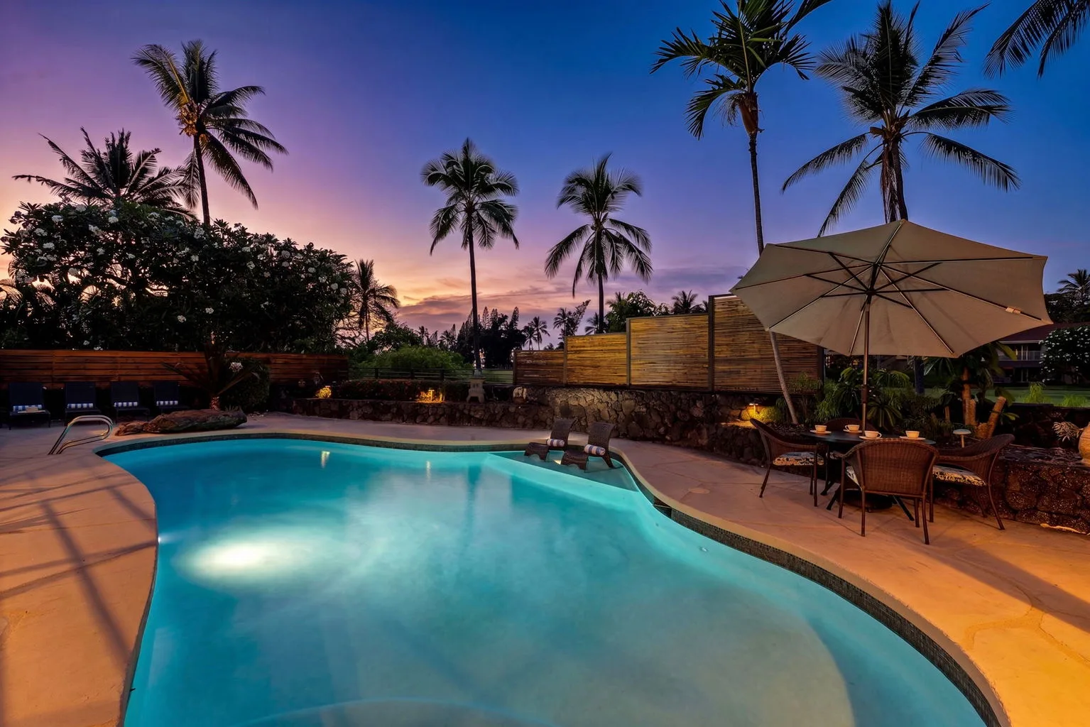 An outdoor swimming pool beside patio seating and palm trees at dusk.