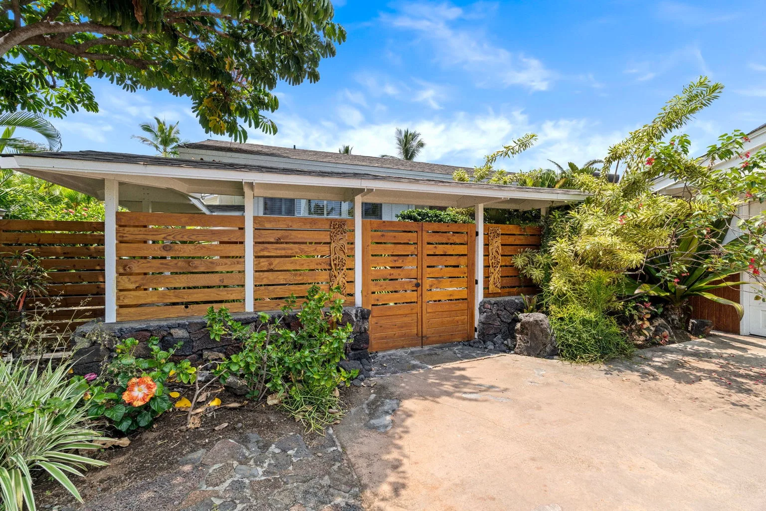 A wooden garden gate and fence sit among tropical plants beside a paved path.
