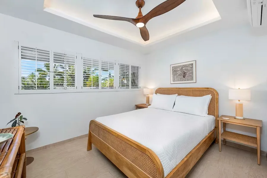 A bright bedroom with a woven bed, bedside tables, and windows with blinds.