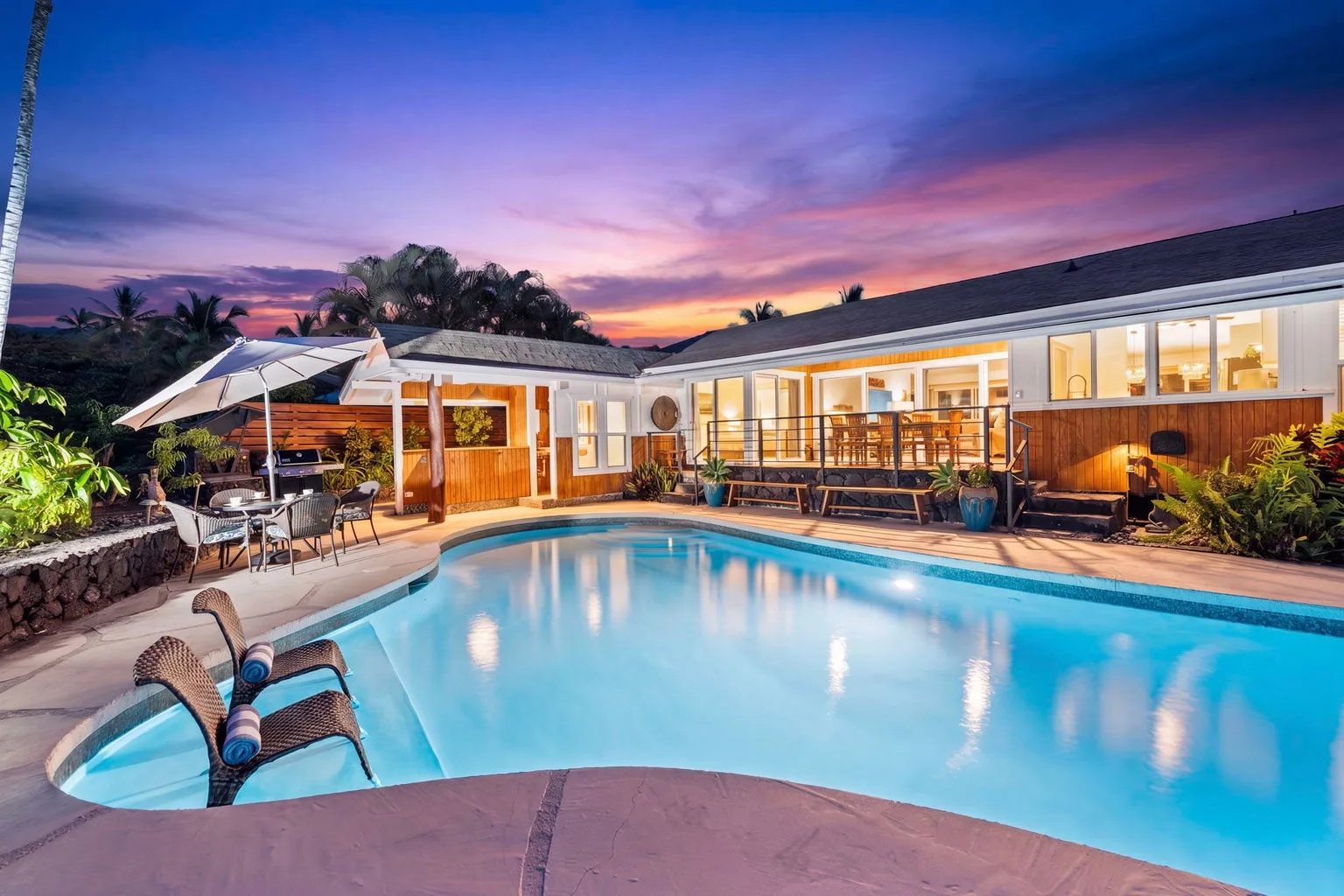 An outdoor swimming pool beside a lit house at sunset.