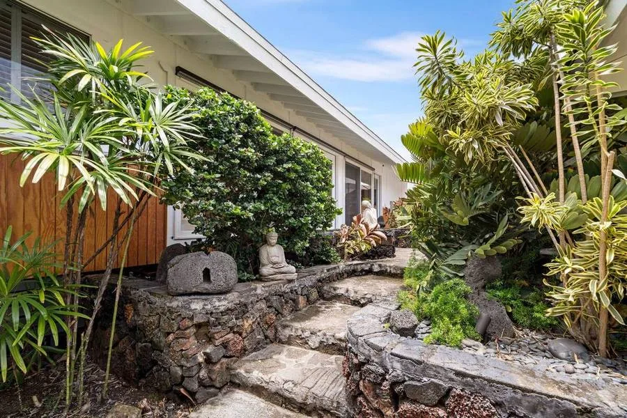 A garden path runs beside a building, with lush plants and a stone statue.