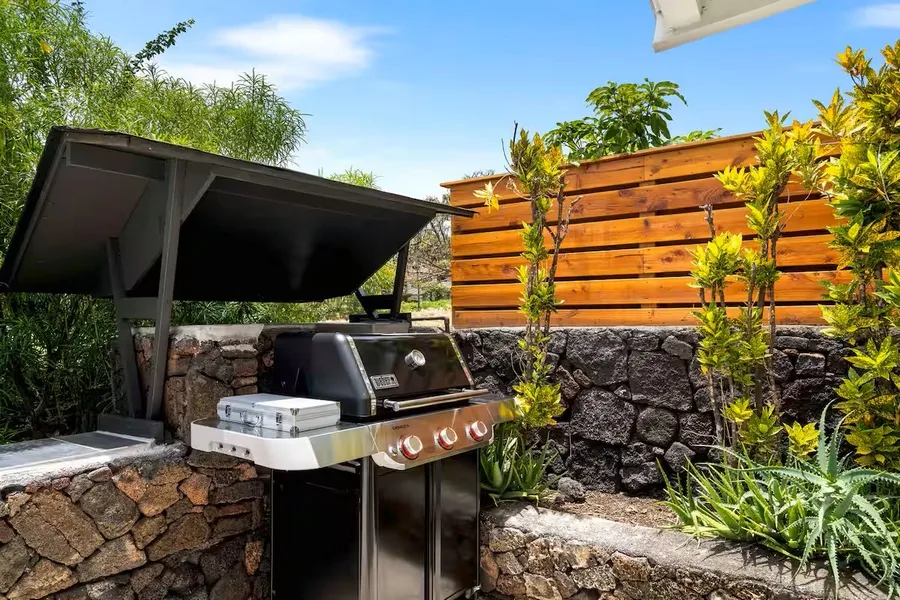 An outdoor grill sits beneath a canopy beside a stone wall and wooden fence.