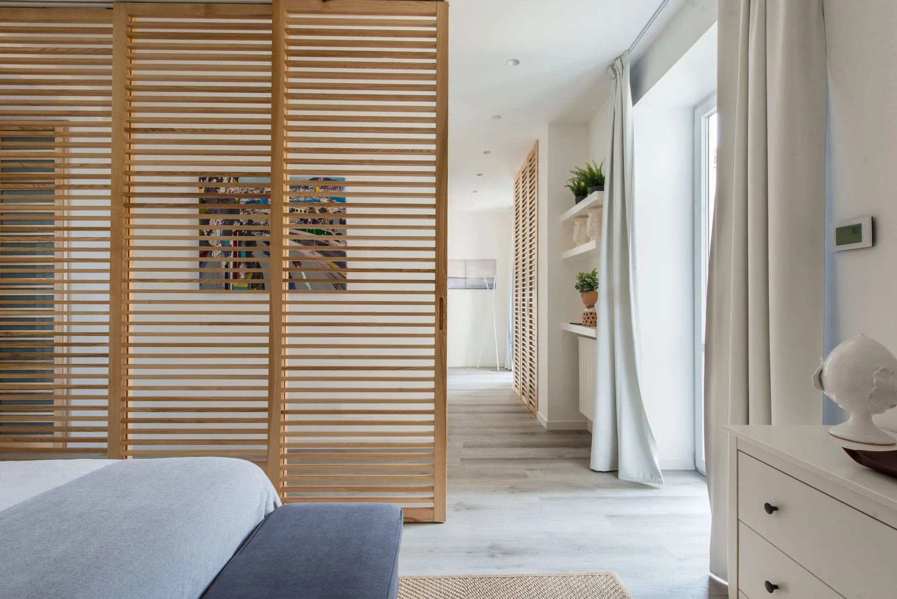 A bright bedroom with a bed, slatted wood partition, and tall curtained windows.