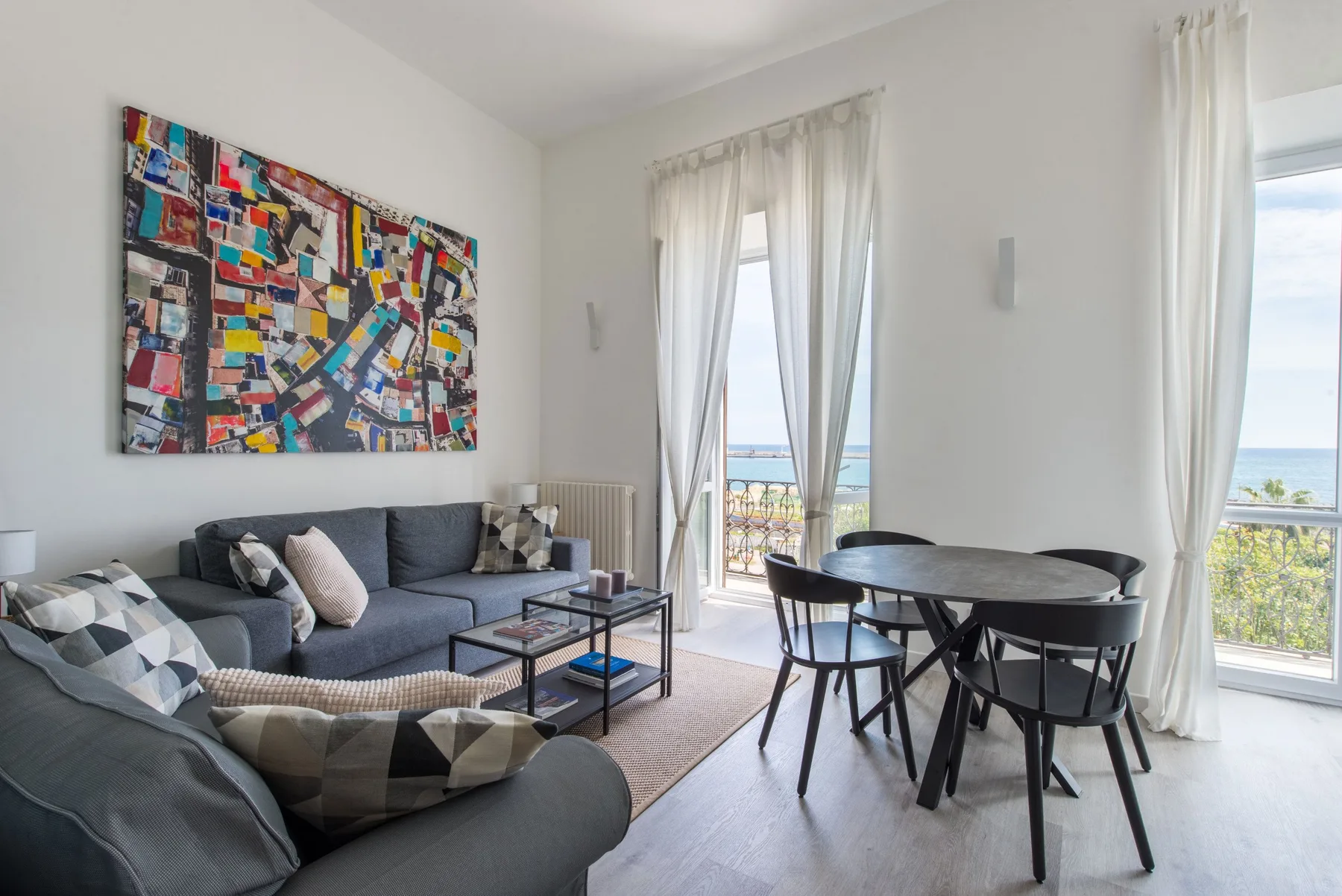 A bright living and dining area with seating, a table, and windows overlooking the sea.