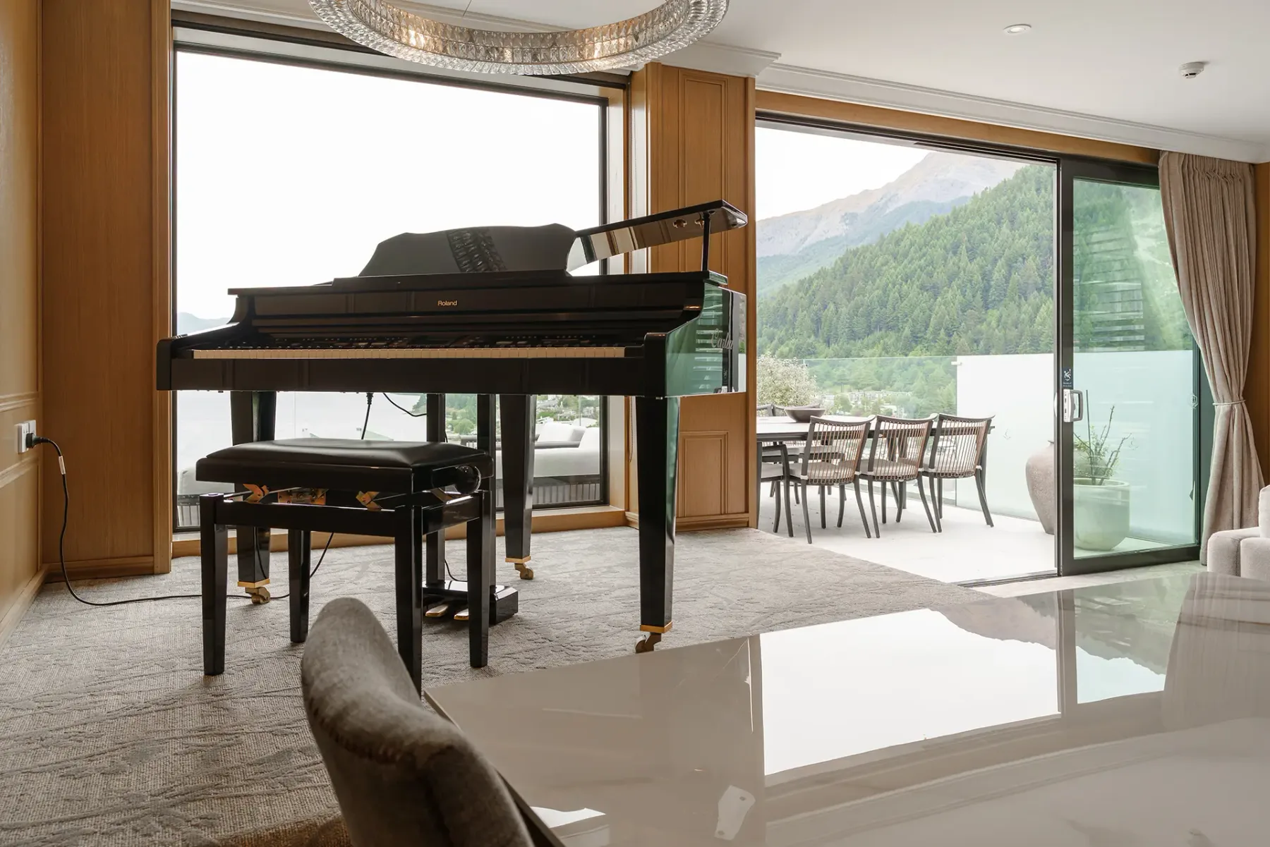 Modern suite room with a grand piano, large windows, and a balcony overlooking forested mountains.