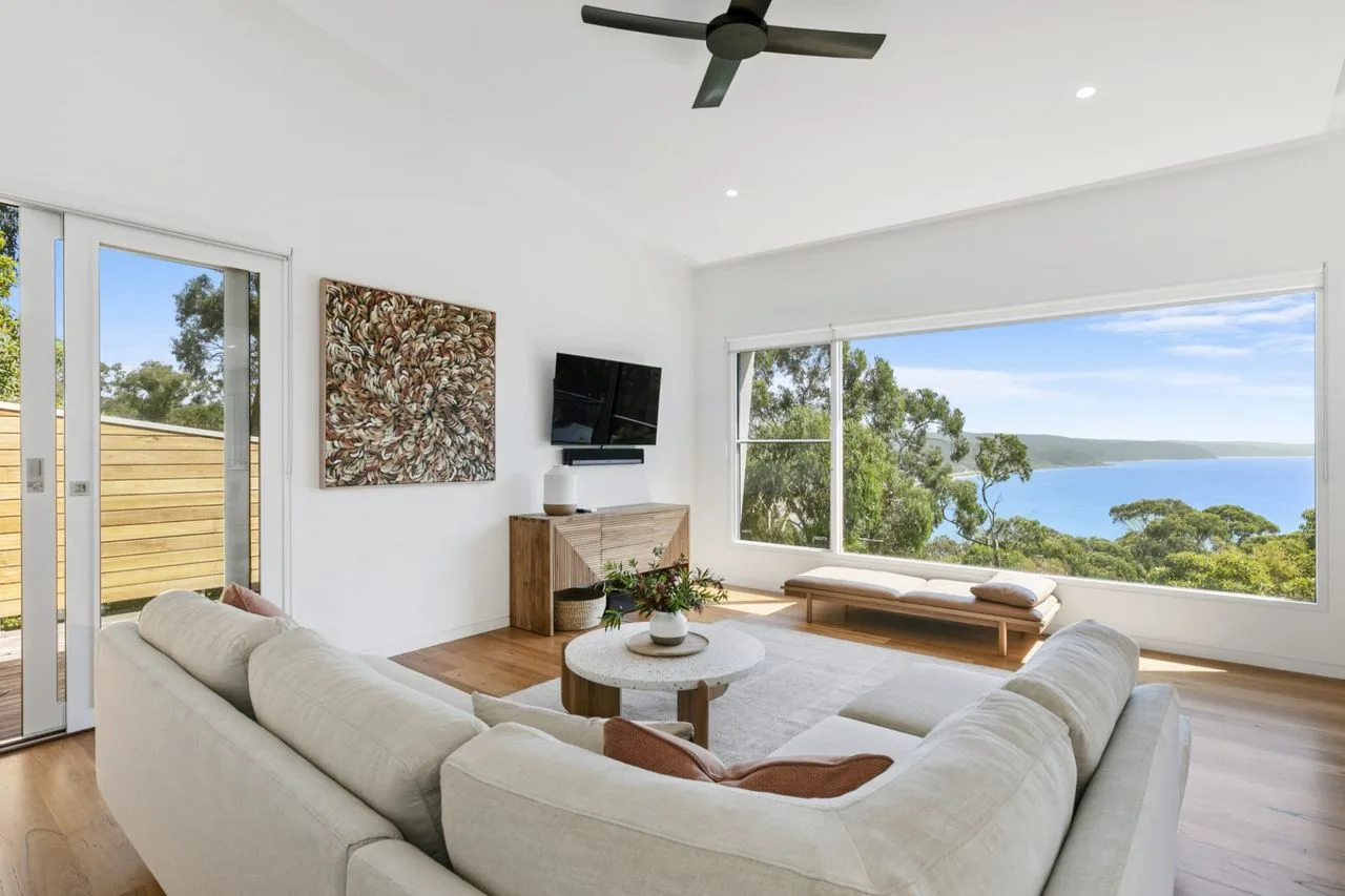 A bright living room has seating beside large windows overlooking trees and water.