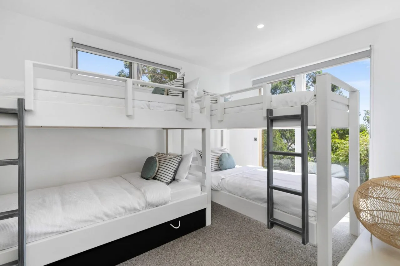 Dormitorio luminoso con literas y otra cama junto a grandes ventanas.